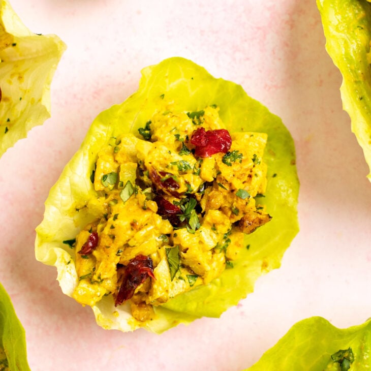 This is a vertical image of lettuce cups on a light pink surface. In the lettuce cups is a yellow curry chicken salad with nuts, herbs, and dried cranberries. The image focuses on one cup in the center with more lettuce cups in the corners of the image.