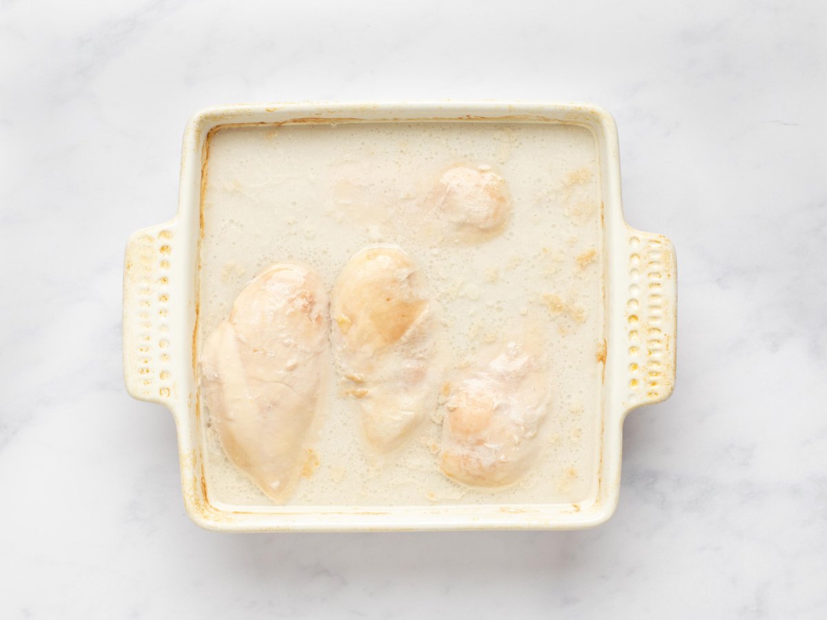 This is an overhead horizontal image of a white ceramic square baking dish. In the dish is cooked chicken in a creamy liquid. The dish sits on a white marble surface.