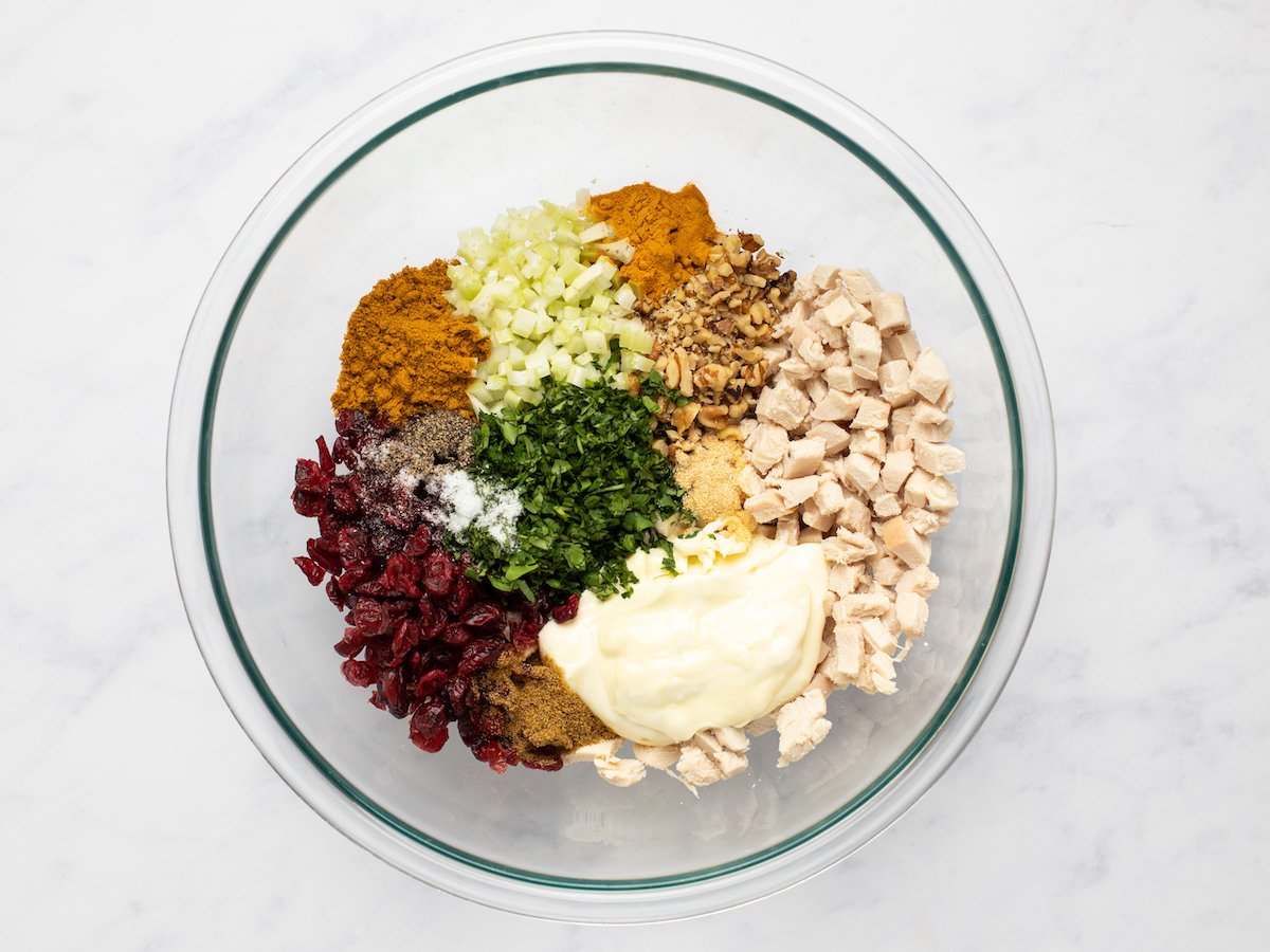 This is an overhead horizontal image of a glass bowl with cubed chicken, chopped walnuts, cilantro, celery, spices, dried cranberries, and mayonnaise in it. The glass bowl sits on a white marble surface.