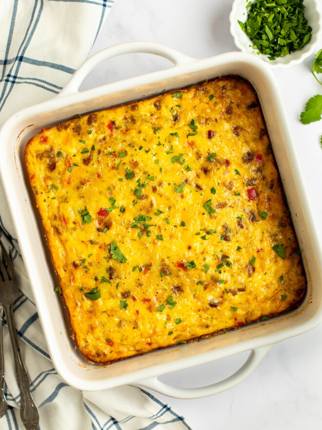 This is an overhead vertical image of a white square casserole dish with handles on the top and bottom. In the dish is a baked egg casserole with pieces of sausage crumbles and pepper peeking through. Chopped herbs are sprinkled on top of the casserole and a small white bowl with more chopped herbs are to the top right corner of the image. The baking dish sits on a white marble surface with a white and blue plaid dish towel to the left side of the image. A couple forks are coming in the bottom left corner of the image.