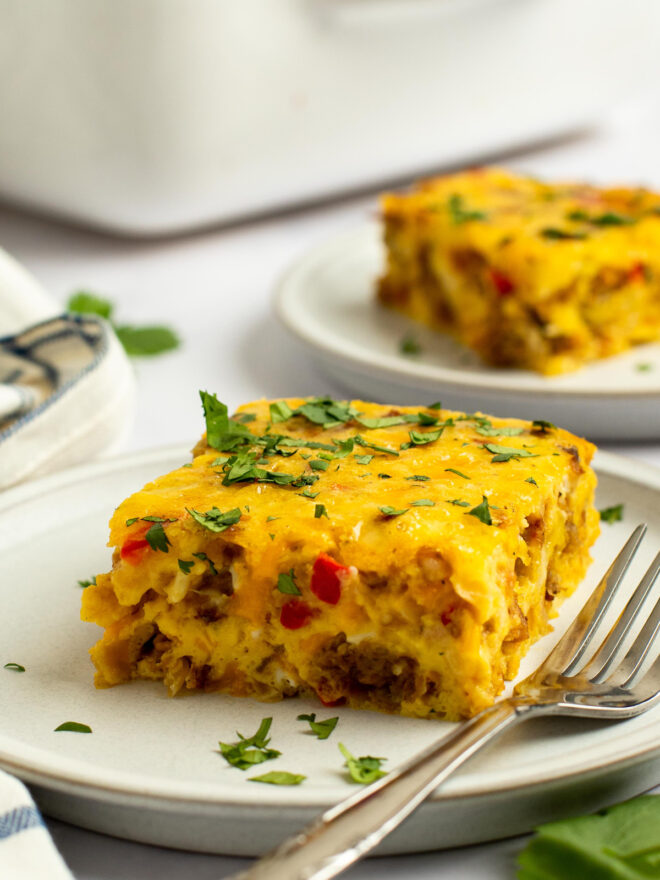 This is a vertical image looking at a piece of an egg casserole from the side. The square of casserole sits on a white plate and is sprinkled with chopped herbs. A fork is coming from the center bottom of the image and rests on the right side of the plate. Another white plate with a slice of egg casserole is blurred in the background toward the top right corner of the image. A white and blue dish towel is on the white marble surface to the left of the front plate. You can see a white baking dish blurred in the far background.