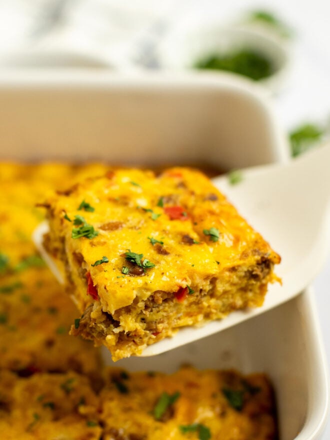 This is a vertical image looking at a piece of egg casserole from the side. A white spatula is lifting the square piece of casserole up from a white baking dish where the rest of the casserole sits. The piece is sprinkled with chopped herbs. The casserole dish is blurred in the background and is sitting on a white surface with more herbs blurred behind it.