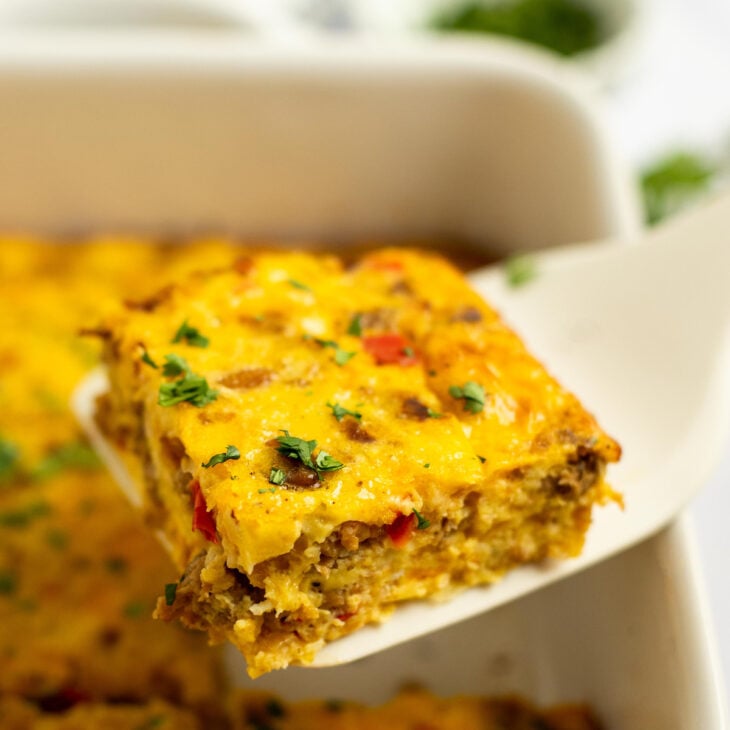 This is a vertical image looking at a piece of egg casserole from the side. A white spatula is lifting the square piece of casserole up from a white baking dish where the rest of the casserole sits. The piece is sprinkled with chopped herbs. The casserole dish is blurred in the background and is sitting on a white surface with more herbs blurred behind it.
