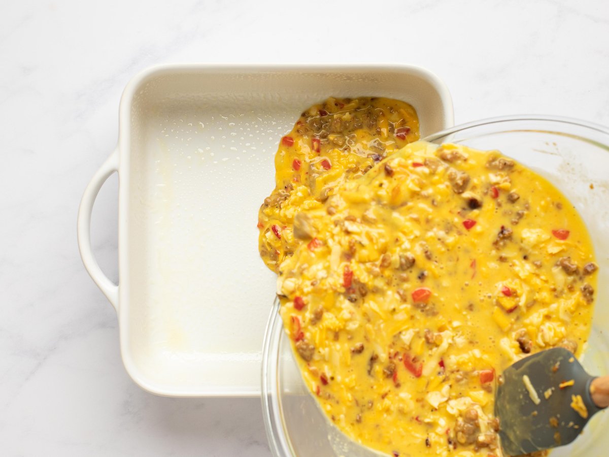 This is an overhead horizontal image of a white square baking dish with handles on either side. A glass bowl filled with an egg, sausage and pepper mixture is coming from the bottom right corner of the image and pouring into the casserole dish. The baking dish sits on a white marble surface.