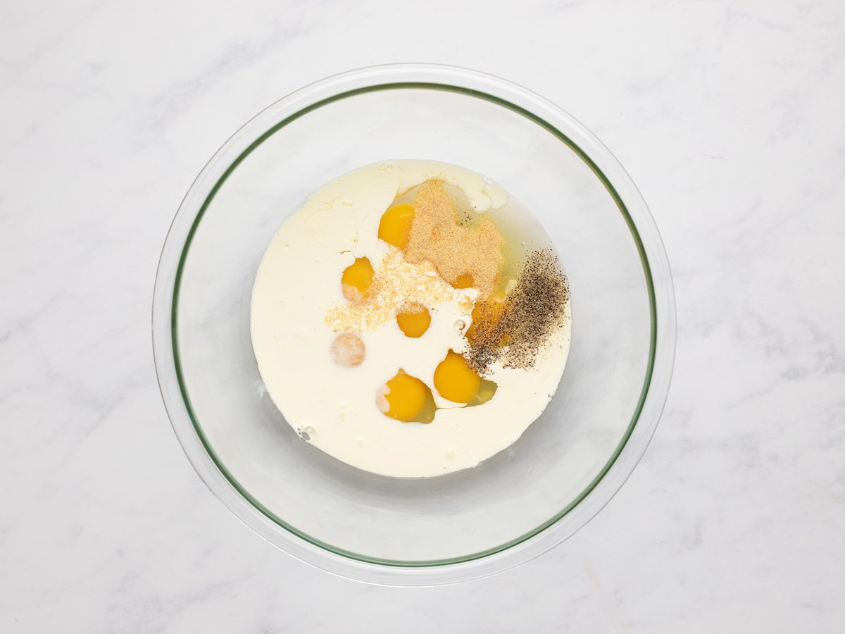 This is an overhead horizontal image of a large glass bowl with eggs, cream, garlic powder, salt, and pepper in it. The bowl sits on a white marble surface.
