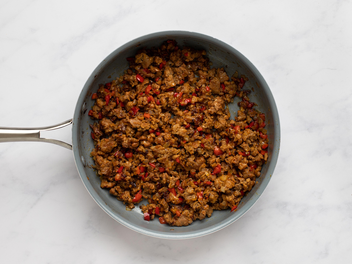 This is an overhead horizontal image of a grey pan with cooked browned sausage crumbles and red pepper bits in it. The handle of the skillet is pointing to the left side of the image. The skillet pan sits on a white marble surface.