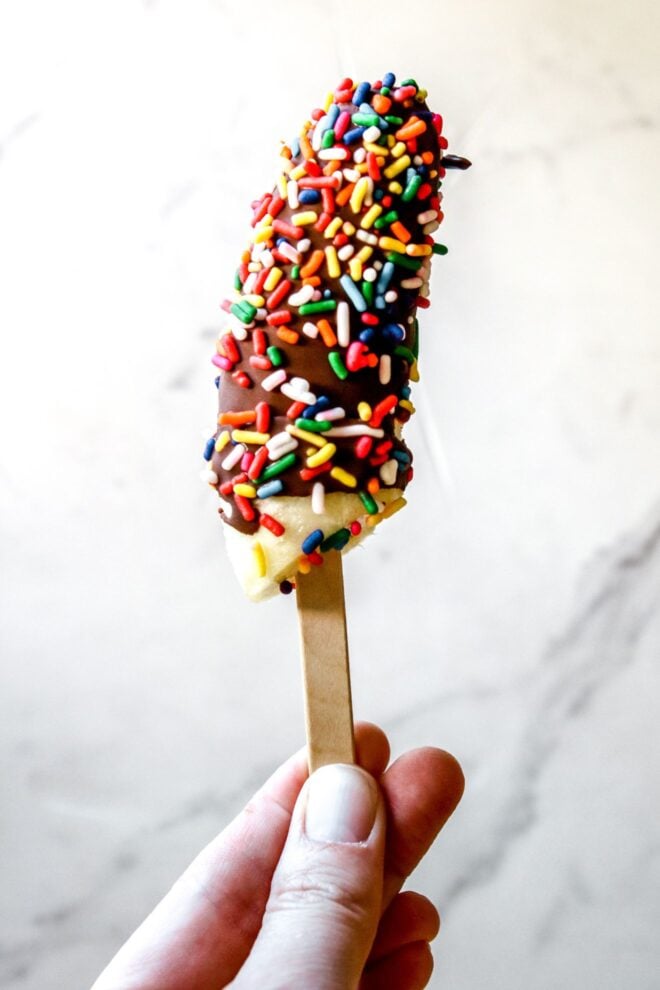 This is a vertical image of a hand holding a banana pop that is coated in hardened chocolate and rainbow sprinkled. The hand is coming up from the bottom holding the popsicle stick with a white marble background.