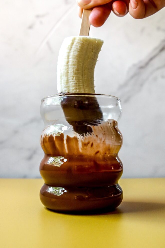 This is a vertical image looking at a clear glass cup from the side. The cup is on a yellow surface with a white marble background. In the cup is melted chocolate. A hand is holding a banana with a popsicle stick and is coming from the top center of the image and appears to have just been inserted into the chocolate cup as there is melted chocolate on the top of the banana and dripping off into the cup.