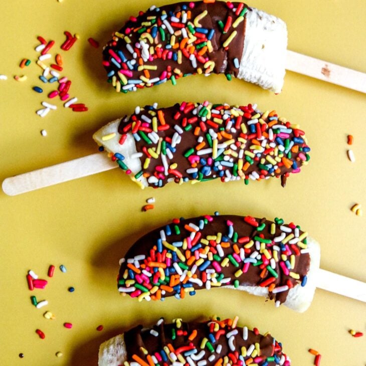 This is a vertical image of banana halves with popsicle sticks inserted in their bottoms. The banana pops are lined up vertically with their popsicle sticks alternating sides. They are coated in hardened chocolate and sprinkled with rainbow sprinkles.