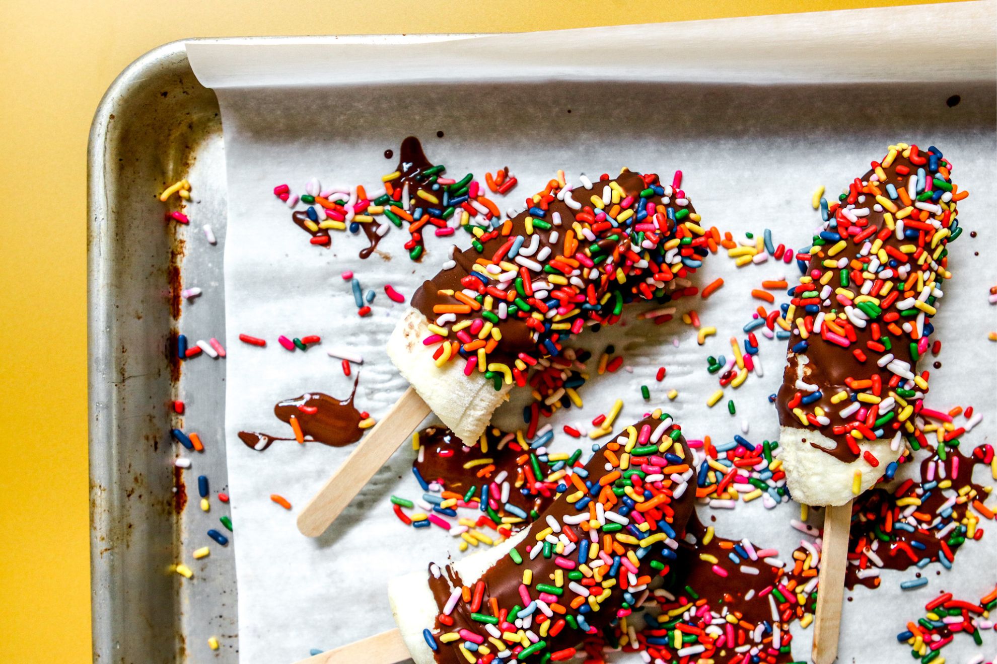 This is an overhead horizontal image of chocolate coated banana pops with rainbow sprinkles. The pops are on a white piece of parchment paper on a baking sheet with lots of melted chocolate and rainbow sprinkles scattered around them. The baking sheet sits on a yellow surface.