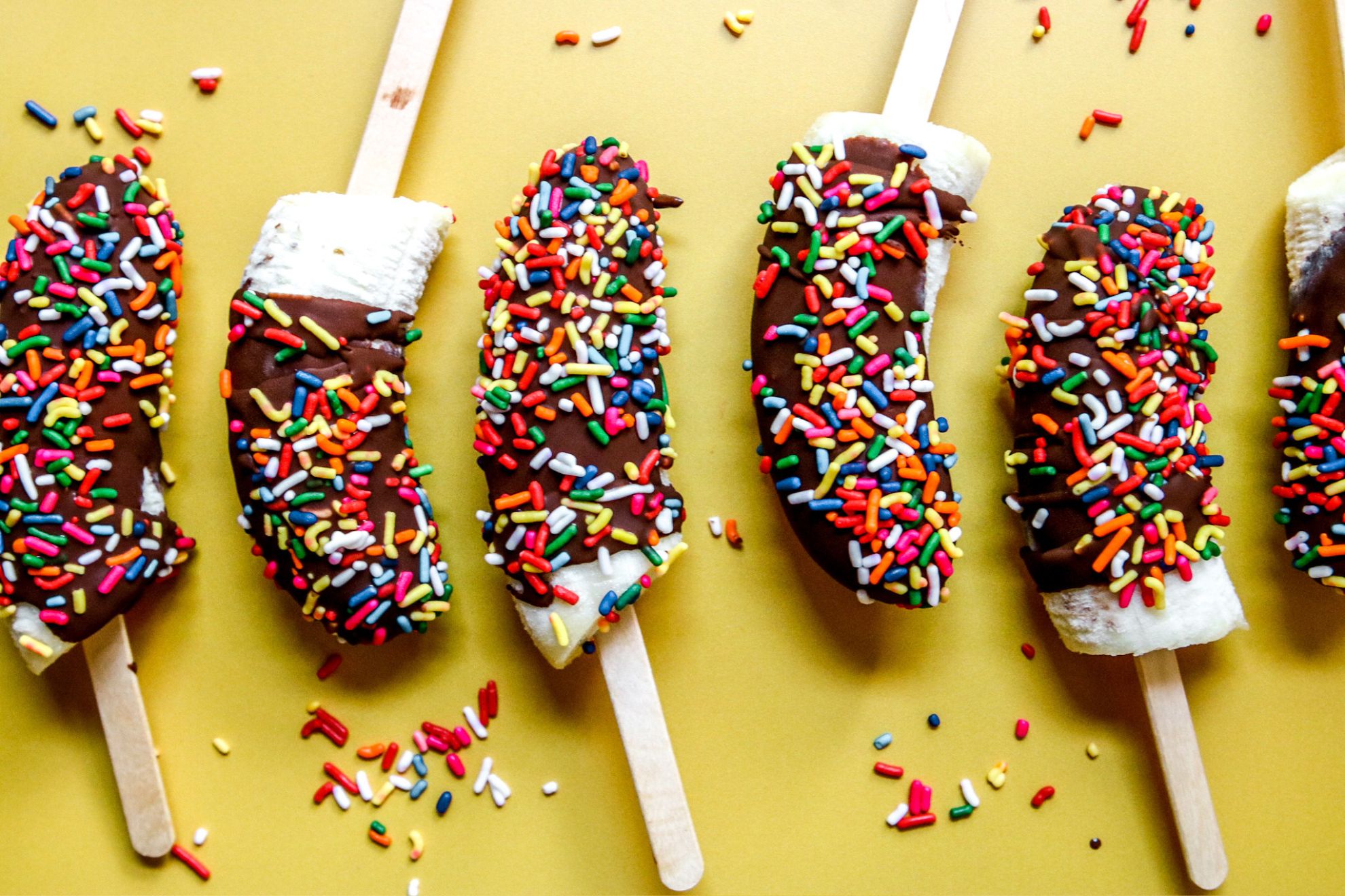 This is a horizontal image of banana halves with popsicle sticks inserted in their bottoms. The banana pops are coated in hardened chocolate and sprinkled with rainbow sprinkles.