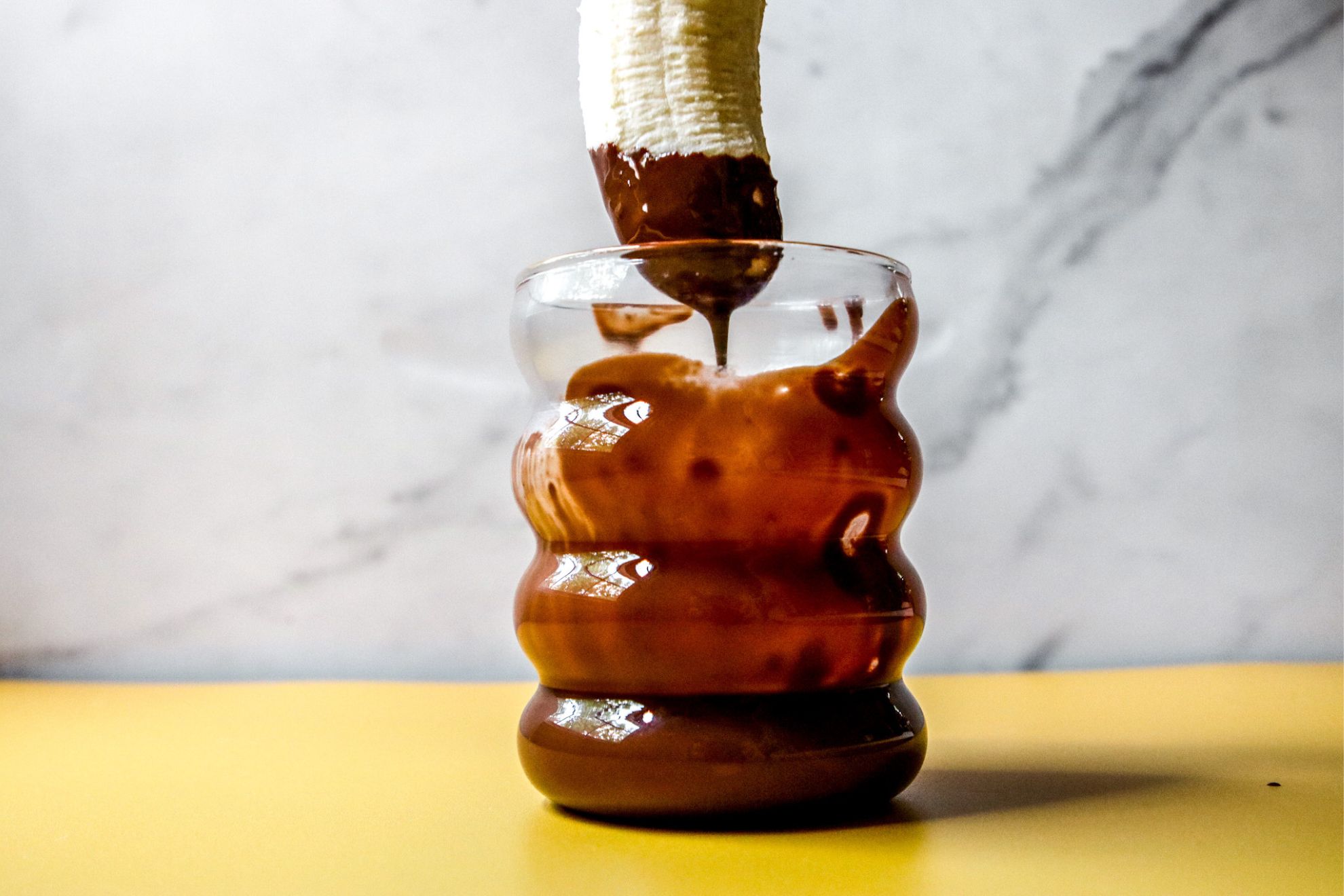 This is a horizontal image looking at a cup from the side. The cup is on a yellow surface with a white marble background. In the cup is melted chocolate. A banana is coming from the top center of the image and appears to have just been inserted into the chocolate cup as there is melted chocolate on the top of the banana and dripping off into the cup.
