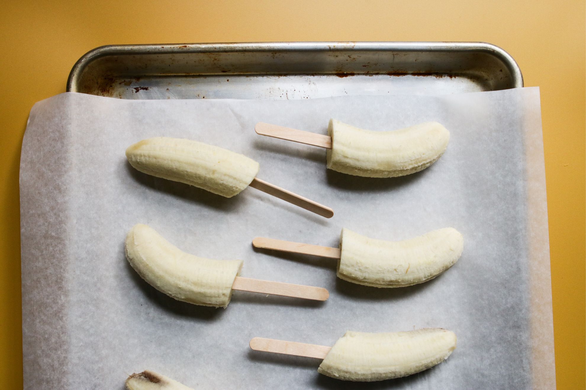 This is an overhead horizontal image of a baking sheet with white parchment paper on a yellow surface. On the parchment paper are banana halves with popsicle sticks inserted into the bottom.