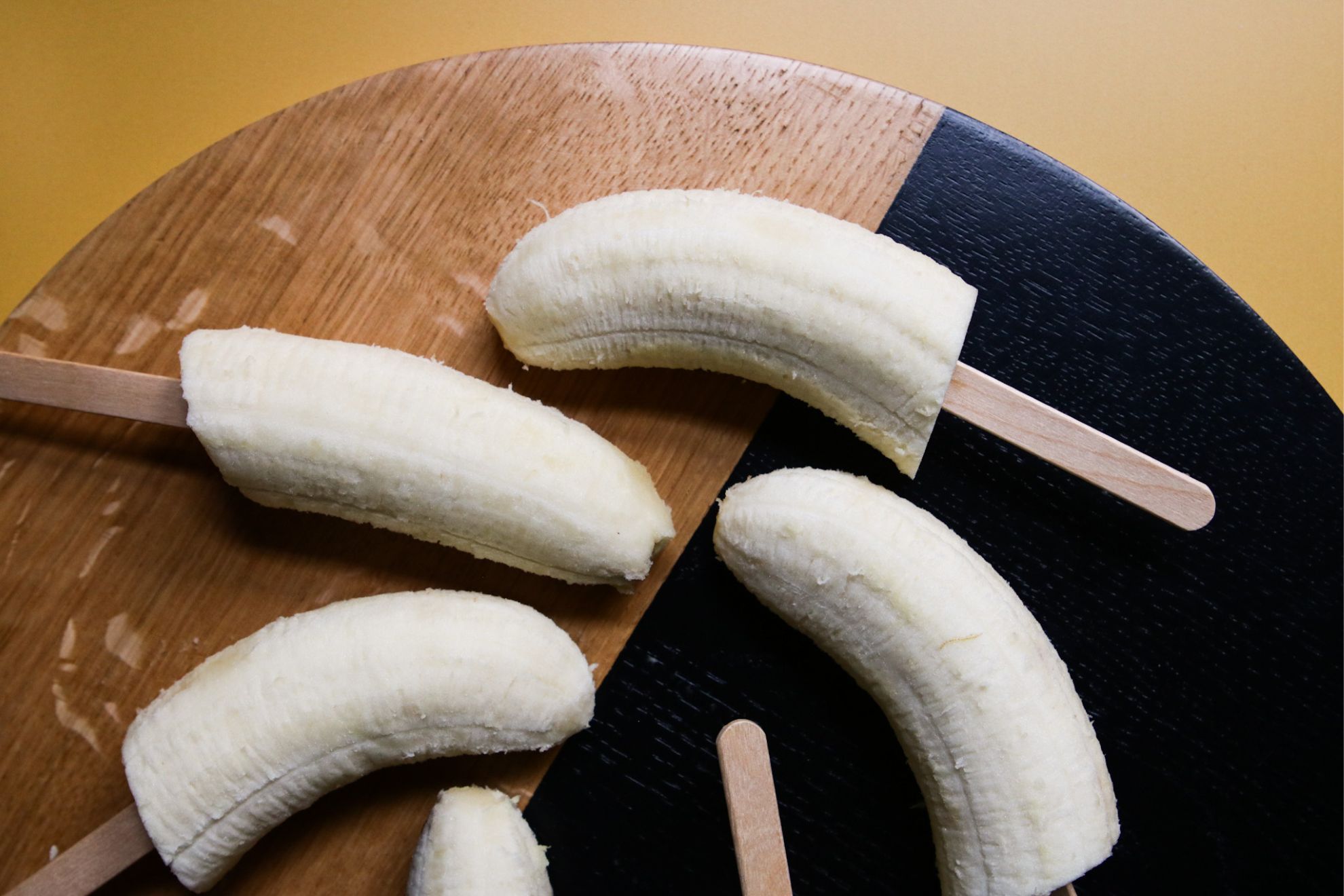 This is an overhead horizontal image of a black and wood circle cutting board on a yellow surface. On the board are banana halves with popsicle sticks shoved in the bottoms.