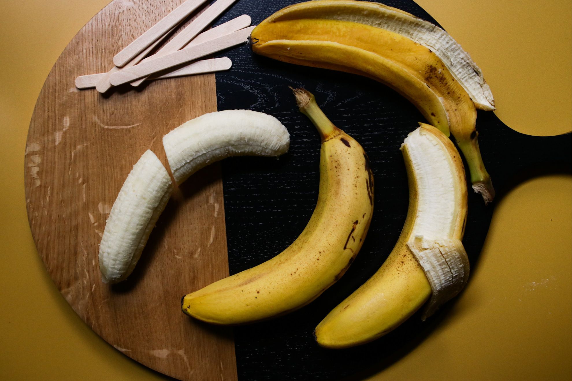 This is an overhead horizontal image of a black and wood circle cutting board on a yellow surface. On the cutting board are three bananas. One is peeled, one is partially peeled and one still has the peel on. Popsicle sticks are also on the cutting board at the top.