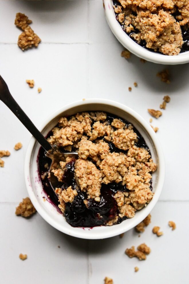 This is a vertical overhead image of one white ramekin with a nutty crumble and blueberry glaze peeking through. A silver spoon is dipping into the contents of the ramekin and leaning against the side with the handle pointing to the top left center of the image. Another ramekin with blueberry crisp is in the top right corner of the image. The ramekins sit on a white square tiled surface with more crumbles that have fallen around them.