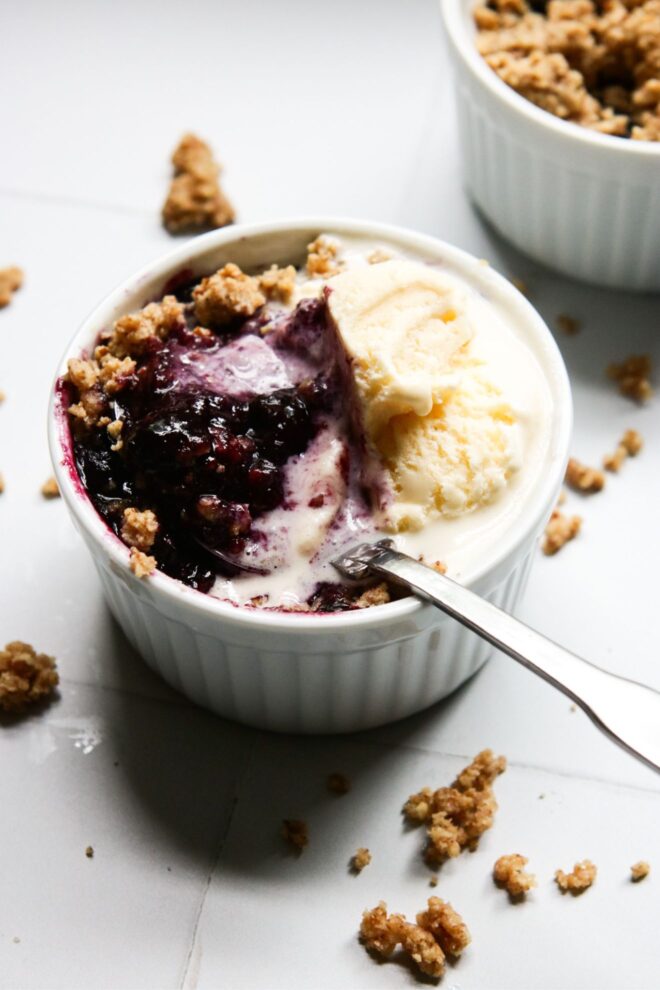 This is a vertical angled view of one white ramekin with a nutty crumble and blueberry glaze scooped up with a silver spoon. The handle of the spoon is sticking out of the ramekin, leaning against the side and pointing at the right side of the image. Vanilla ice cream is on top of the crumble, mainly to the top right of the ramekin, and melting into it. Another ramekin with blueberry crisp is in the top right corner of the image. The ramekins sit on a white square tiled surface with more crumbles that have fallen around them.