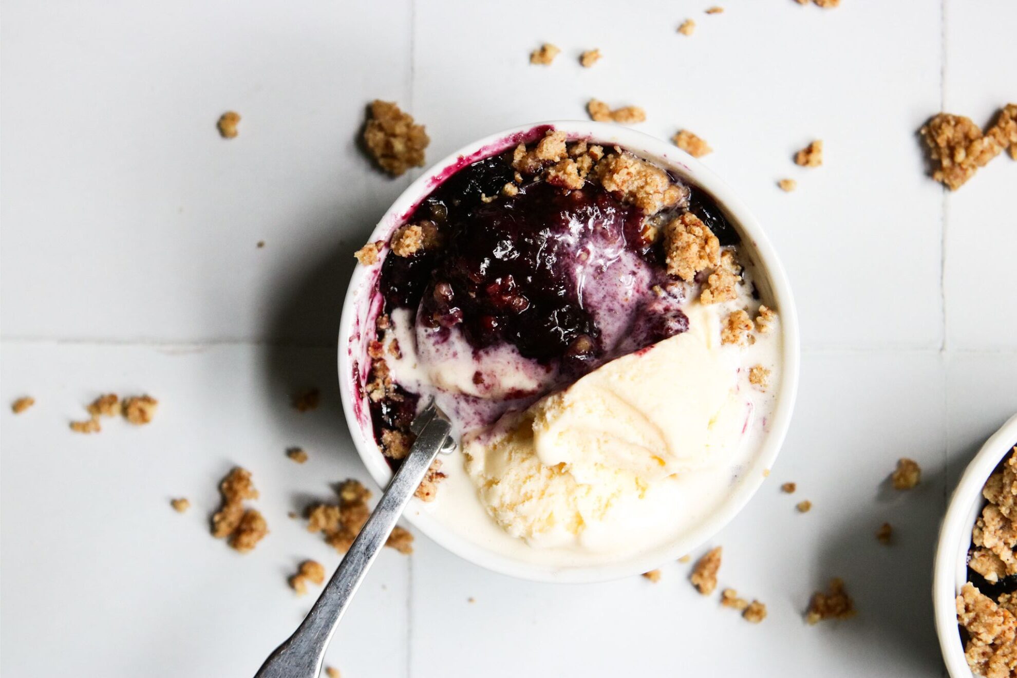 This is a vertical overhead image of one white ramekin with a nutty crumble and blueberry glaze scooped up with a silver spoon. The handle of the spoon is sticking out of the ramekin, leaning against the side and pointing at the right side of the image. Vanilla ice cream is on top of the crumble, mainly to the top right of the ramekin, and melting into it. Another ramekin with blueberry crisp is in the top right corner of the image. The ramekins sit on a white square tiled surface with more crumbles that have fallen around them.