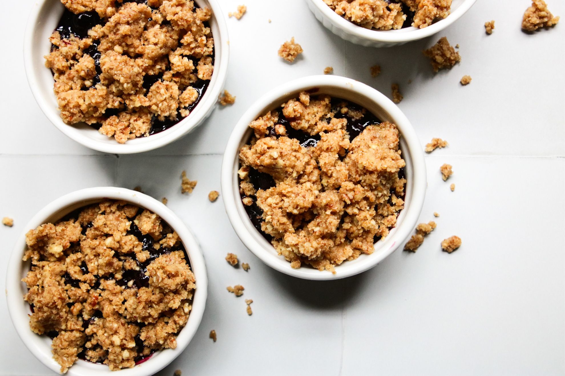 This is an overhead horizontal image of four small white ramekins with a nutty crumble on top. Peeking through the nutty crisp is a blueberry glaze. The ramekins sit on a white square tiled surface with more crumbles that have fallen around them.