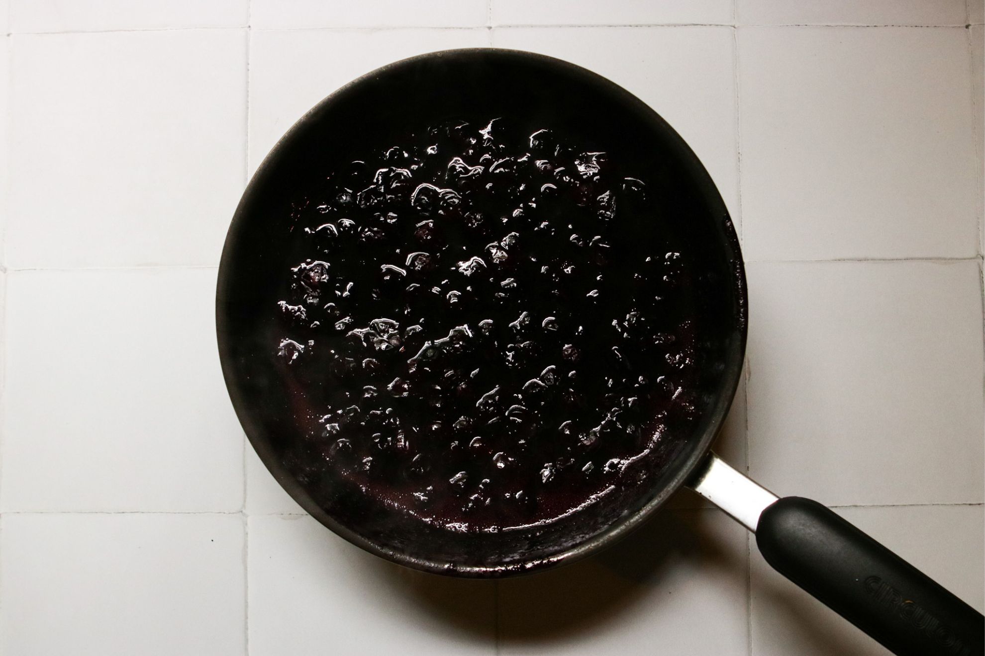 This is an overhead horizontal image of a small saucepan with a reduced berry glaze in it. The handle of the pan is pointing to the bottom right corner of the image. The pan sits on a white square tile surface.
