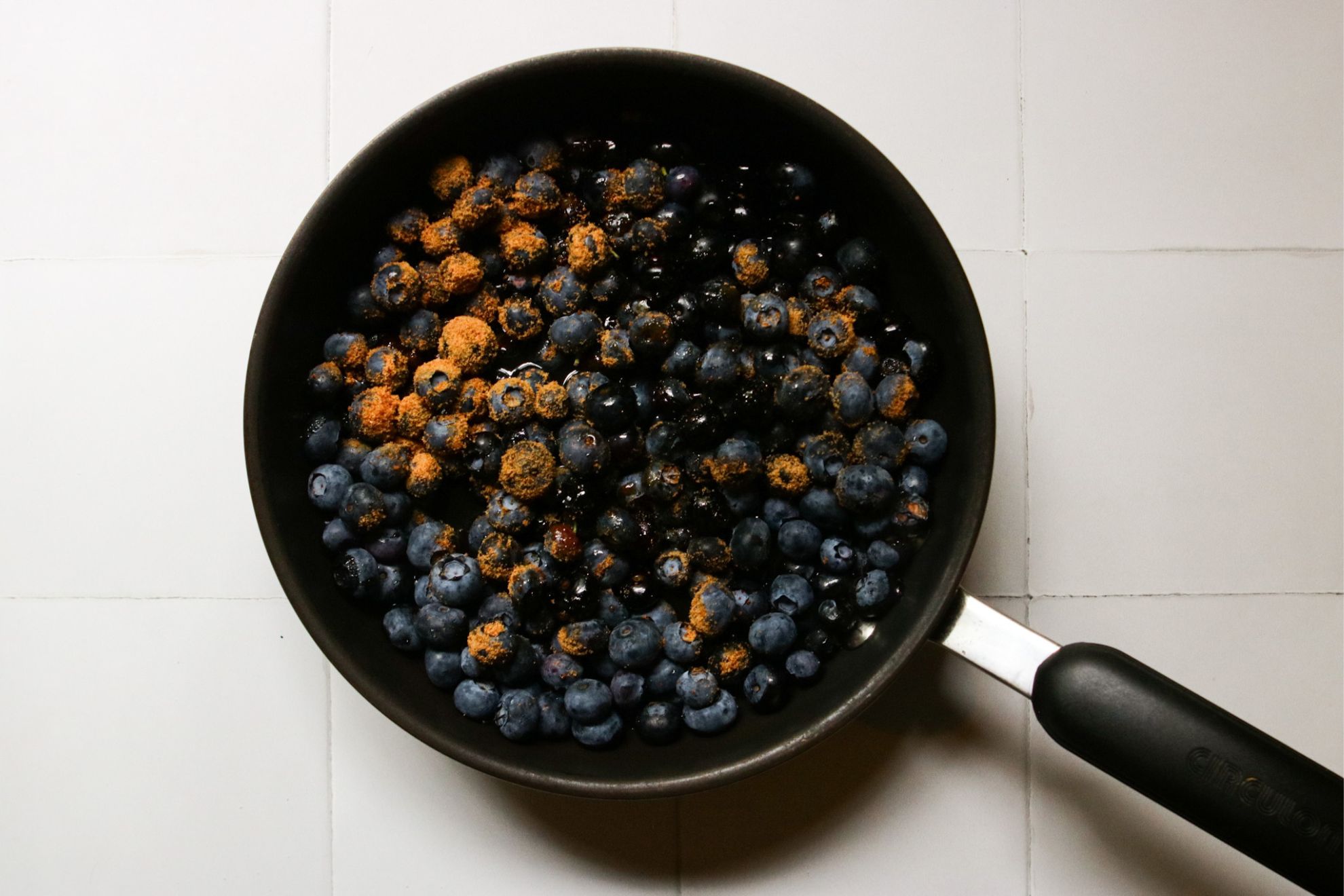 This is an overhead horizontal image of a small saucepan with blueberries and coconut sugar. Some of the blueberries in the pan appear to be wet. The handle of the pan is pointing to the bottom right corner of the image. The pan sits on a white square tile surface.