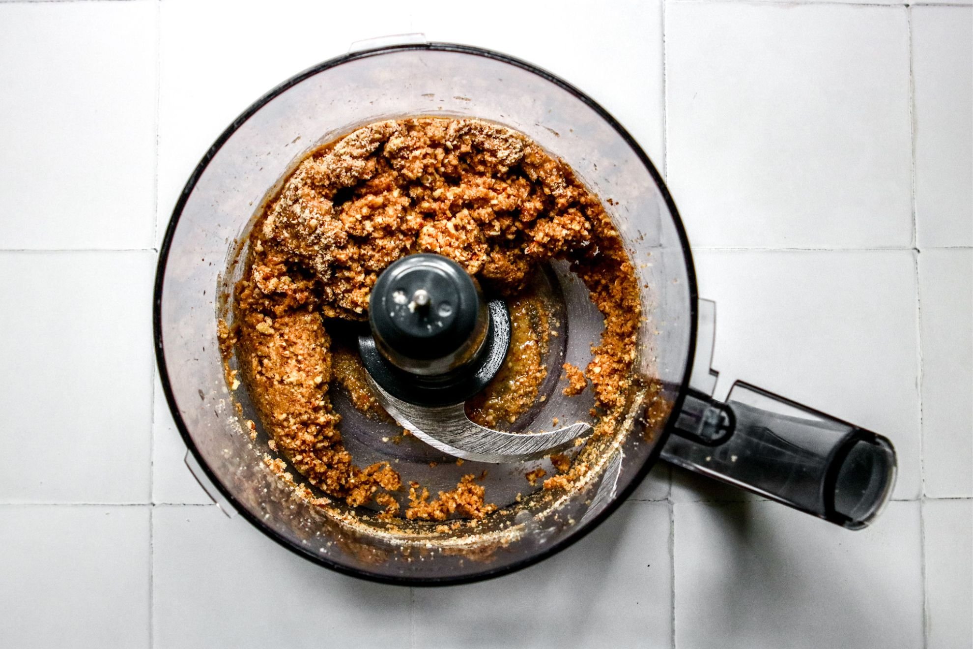 This is an overhead horizontal image of a food processor with a wet, crumbly nutty mixture. The clear plastic food processor bowl sits on a white square tile surface. The handle of the food processor is pointing to the right bottom corner of the image.