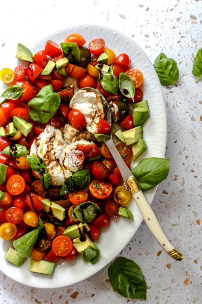This is an overhead vertical image of a white oval plate on a terrazzo surface. On the plate is a caprese salad. The image focuses on a ball of burrata pulled apart in the center of tomatoes cut in half, chunks of avocado, and basil leaves. The salad is drizzled with olive oil and balsamic vinegar. A silver spoon is scooping up some burrata and tomatoes and leaning on the plate.