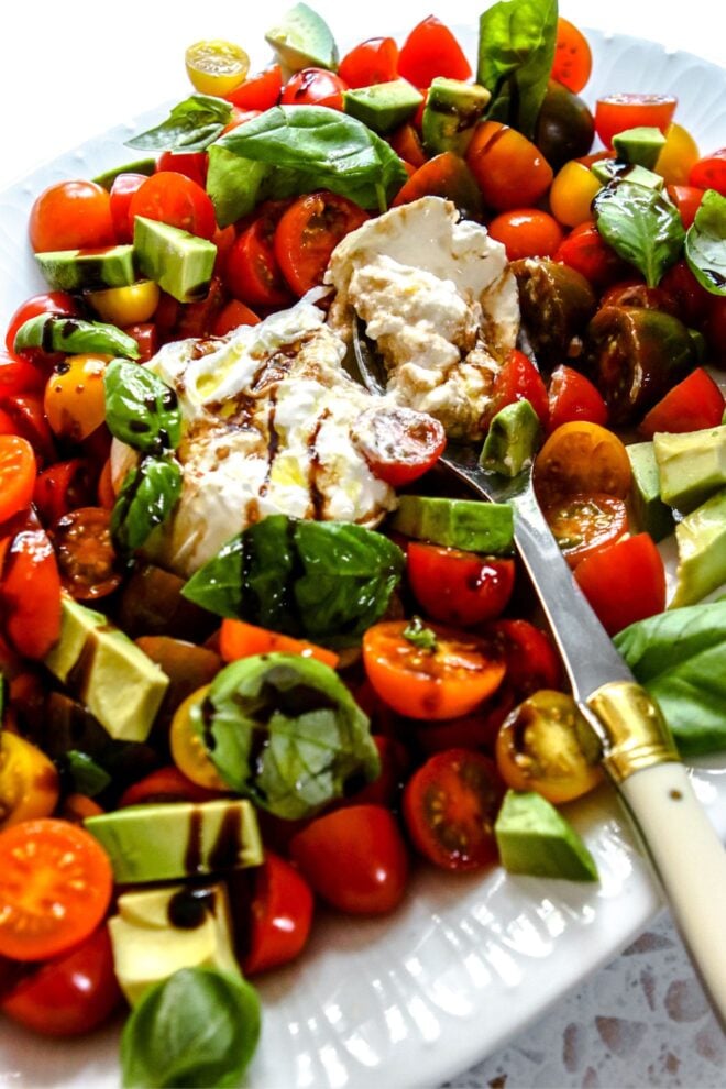 This is a vertical image looking at a caprese salad from the side. The image focuses on a ball of burrata pulled apart. Around the burrata are small cherry tomatoes cut in half, chunks of avocado, basil leaves and a drizzle of olive oil and balsamic vinegar. A silver spoon is leaning against the side of the oval plate and scooping up some burrata.