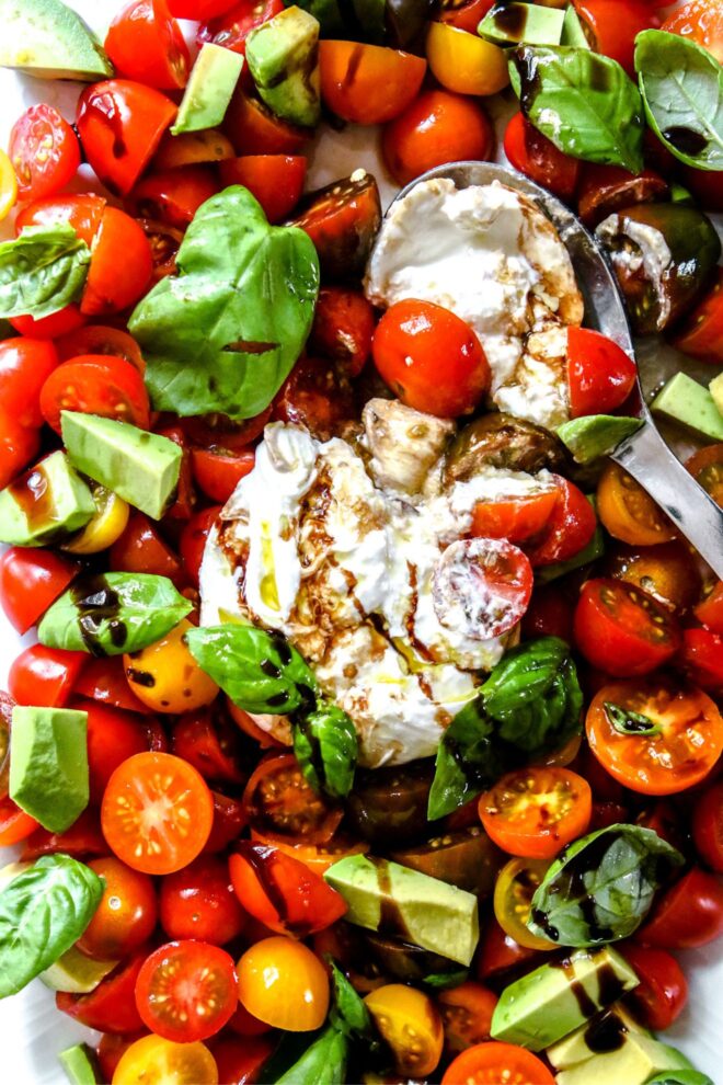 This is an overhead vertical image of a closeup of a caprese salad. The image focuses on a ball of burrata pulled apart in the center of tomatoes cut in half, chunks of avocado, and basil leaves. The salad is drizzled with olive oil and balsamic vinegar. A silver spoon is scooping up some burrata and tomatoes and leaning on the plate.