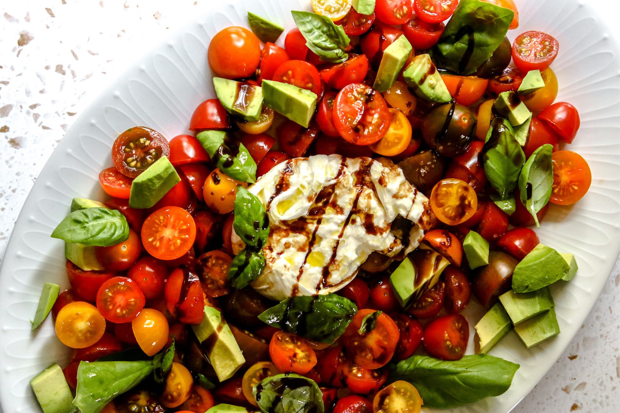 This is an overhead horizontal image of a large white oval plate on a light terrazzo surface. One the plate are small cherry tomatoes cut in half with chunks of avocado scattered around and a ball of burrata pulled apart in the center. Basil leaves are scattered on top of the caprese salad and everything is drizzled with olive oil and balsamic vinegar.