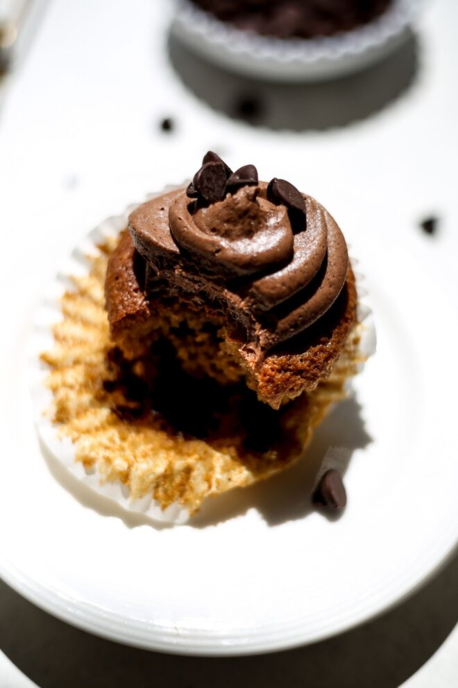 This is a vertical image looking at a cupcake from an angled perspective. The cupcake is a caramel color with chocolate swirled frosting on top and some chocolate chips. A white cupcake liner is pulled away from the cupcake and a bite is taken out of the cupcake. The cupcake sits on a white plate on a white surface. Blurred in the background is a small white bowl of chocolate chips with a few chocolate chips scattered around it.