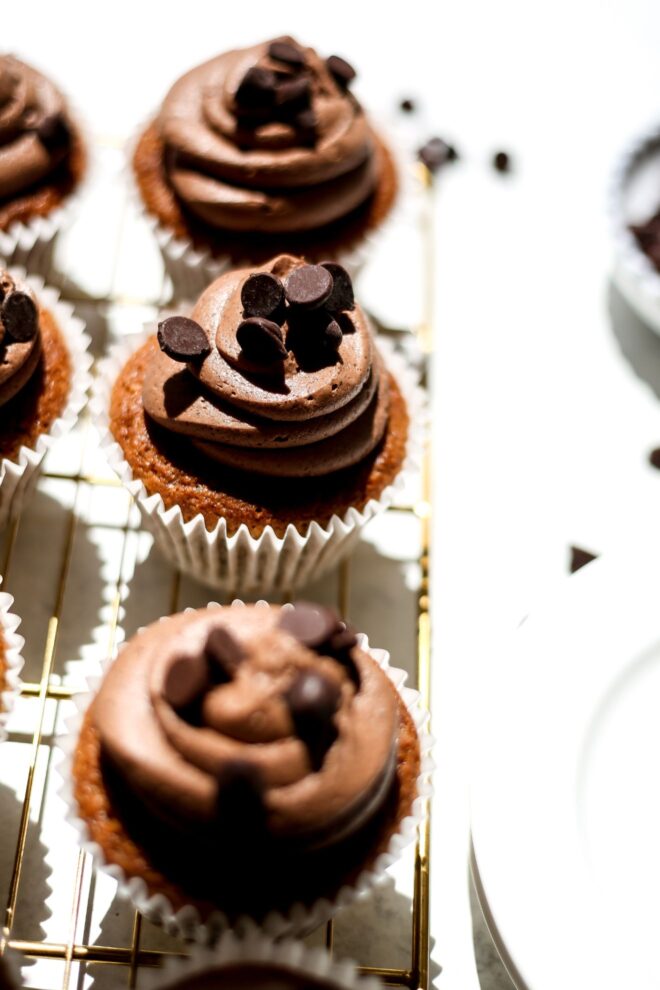This is a vertical image looking at a line of three cupcakes from a side angle perspective. The image focuses on the center cupcake that is a caramel color with swirled chocolate frosting on top with chocolate chips. The line of cupcakes sits on a gold cooling rack on a white surface. Blurred to the right of the cooling rack is a small bowl with chocolate chips and more chocolate chips scattered around it.