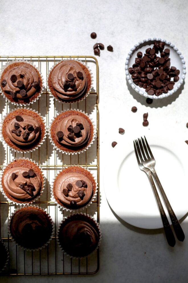 This is an overhead vertical image of a gold cooling rack on a white surface coming from the left of the image. On the cooling rack are caramel colored cupcakes with white cupcake liners. Most of the cupcakes have chocolate frosting swirled on top with chocolate chips on top. To the top right corner of the image is a a small white bowl with chocolate chips and some more chocolate chips scattered around it. Beneath the small bowl is a white plate with two forks lying on top.