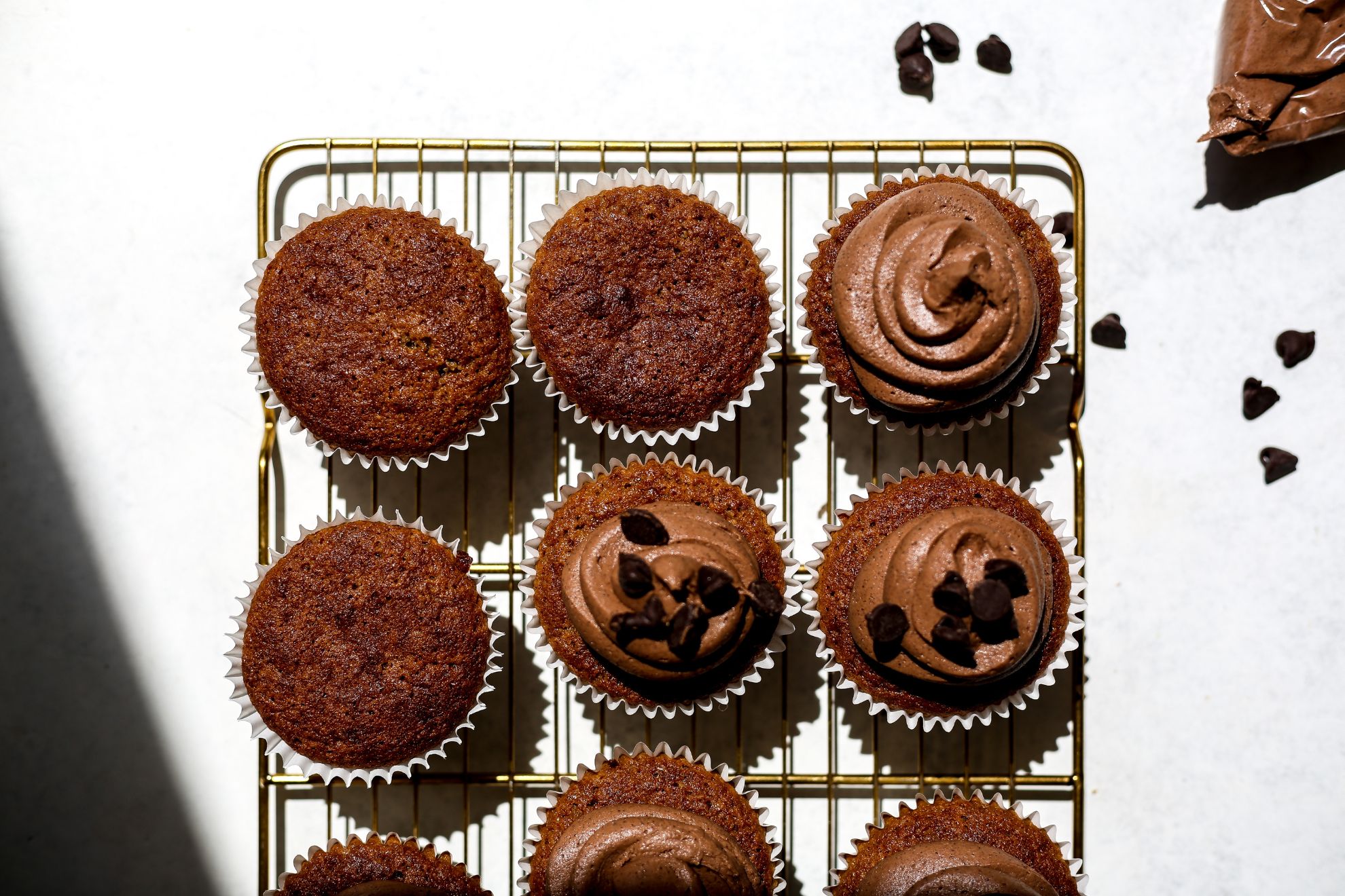 This is an overhead horizontal image of a gold cooling rack on a white surface. On the cooling rack are caramel colored cupcakes with white cupcake liners. Half of the cupcakes have chocolate frosting swirled on top with chocolate chips on top. To the top right corner of the image is a plastic baggie with chocolate frosting and some chocolate chips scattered around it.