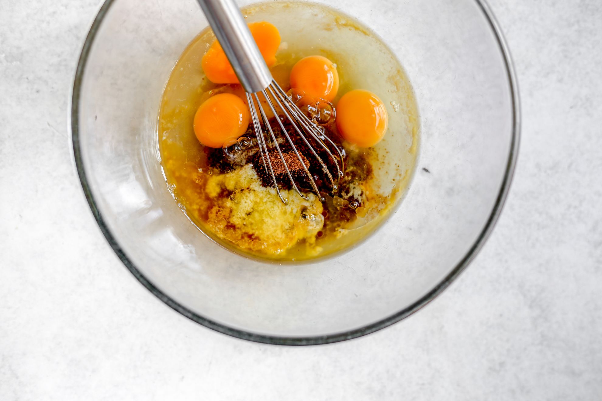 This is an overhead horizontal image of a glass bowl with eggs, coconut sugar and more wet ingredients in it. A whisk is in the center of the bowl with the handle leaning against the side pointing to the top center of the image. The ingredients aren't yet mixed together. The bowl sits on a light grey/white surface.