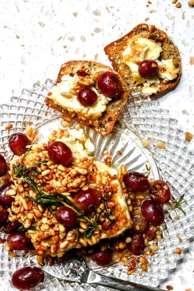 This is a vertical overhead image of a block of feta crusted with toasted pine nuts. Blistered red grapes are on top of the feta and around it on the beveled glass plate. A sprig of rosemary is on top of the feta. A silver cheese knife is leaning on the bottom part of the plate with the handle pointing to the right corner of the image. Nutty crackers with dried fruit in them lay on the white terrazzo surface to the top center of the image. On the crackers is some feta spread out, a couple grapes and toasted pine nuts.