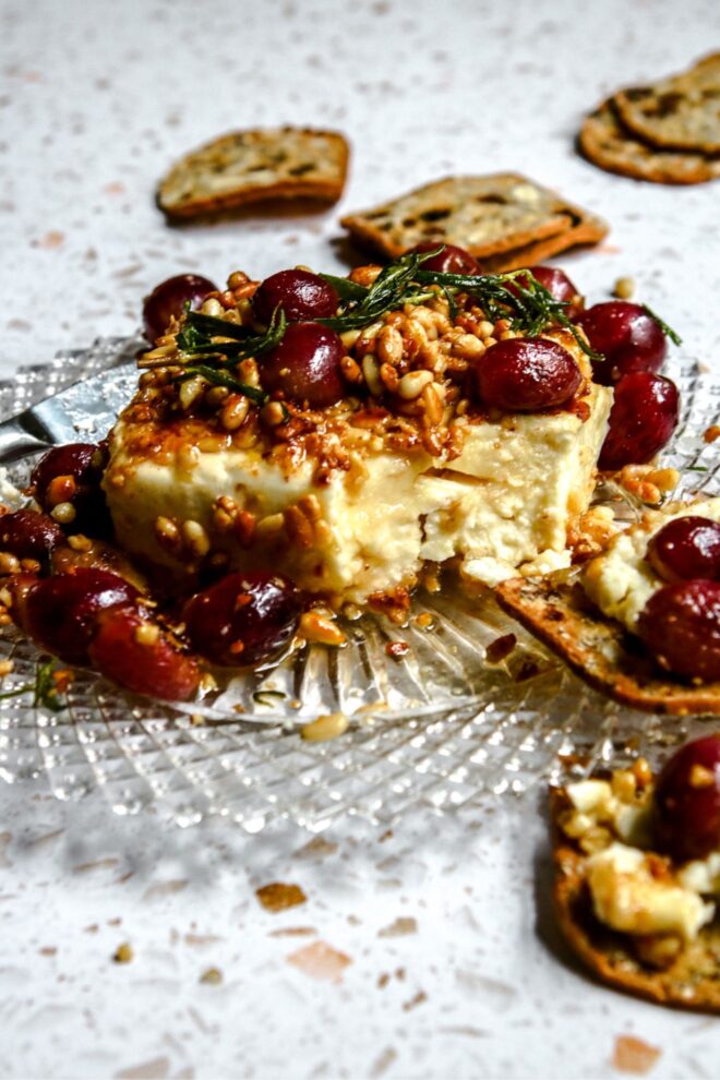 This is a vertical image looking at a block of feta from the side. The feta block is coated with toasted pine nuts. Blistered red grapes are on top of the feta and around it on the beveled glass plate. A sprig of rosemary is on top of the feta. Nutty crackers with dried fruit in them lay on the white terrazzo surface in front of and behind the plate.