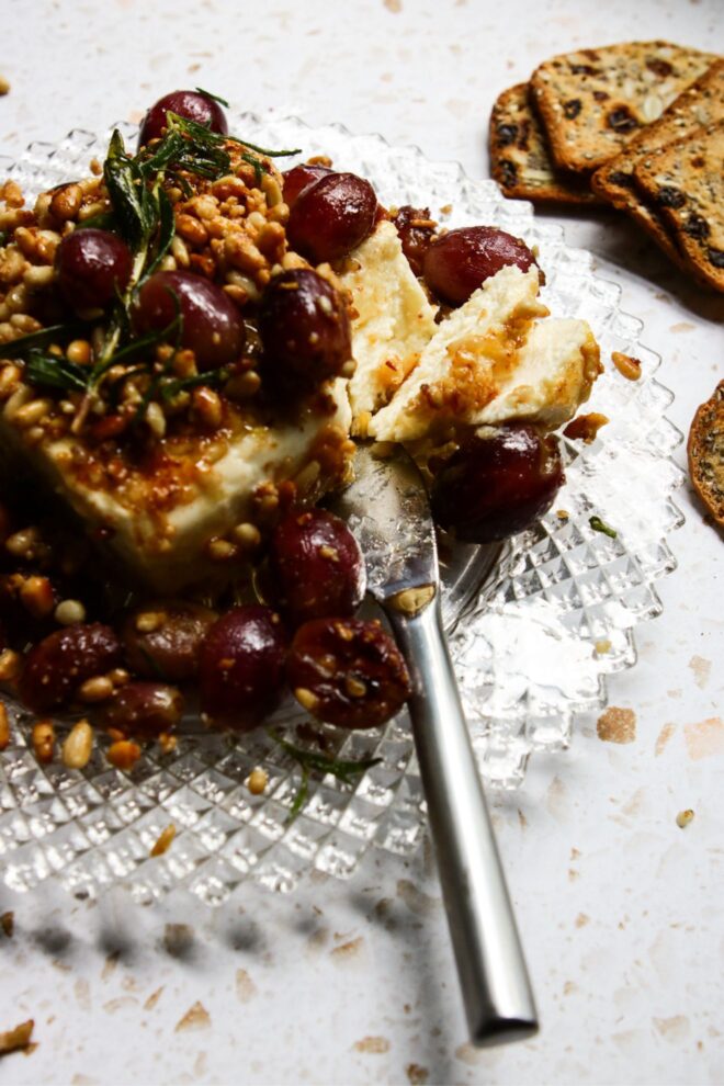 This is a vertical image looking at a block of feta from the side. The feta block is coated with toasted pine nuts. Blistered red grapes are on top of the feta and around it on the beveled glass plate. A sprig of rosemary is on top of the feta. A silver cheese knife sliced a few pieces of feta and is leaning on the side of the plate with the handle pointing to the bottom center edge of the image, toward the viewer. Nutty crackers with dried fruit in them lay on the white terrazzo surface.