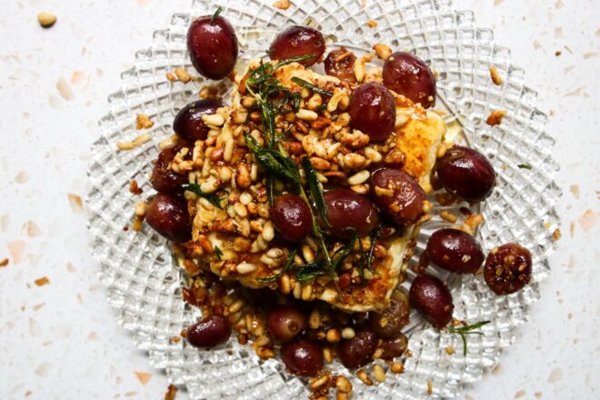 This is an overhead horizontal image of a beveled glass plate with a block of feta on it. The feta is crusted with toasted pine nuts and topped with a sprig of rosemary. Blistered grapes are on the feta and around it. The plate sits on a white and tan terrazzo surface.
