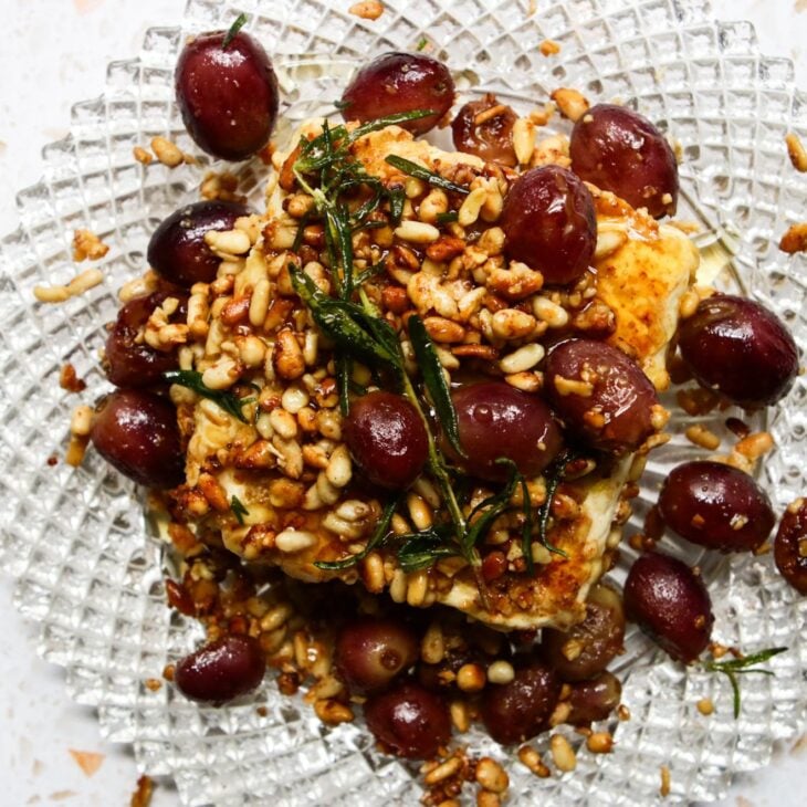 This is an overhead horizontal image of a beveled glass plate with a block of feta on it. The feta is crusted with toasted pine nuts and topped with a sprig of rosemary. Blistered grapes are on the feta and around it. The plate sits on a white and tan terrazzo surface.