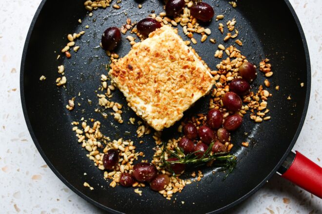 This is an overhead horizontal image of a block of pine nut crusted feta. The feta block sits in a large black skillet with a red handle pointing to the bottom right corner of the image. The pine nuts are toasted light brown, about 1 cup of red grapes and a sprig of rosemary are also in the pan. More crushed pine nuts are around the block of feta.