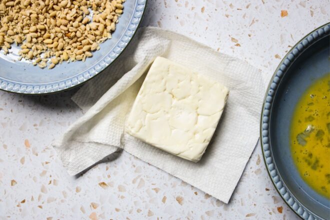 This is an overhead horizontal image of a block of feta on a paper towel in the middle. To the top left corner is a shallow light blue bowl of crushed pine nuts. To the bottom right corner is a shallow blue bowl with a whisked egg. The items are on a white and tan terrazzo surface.