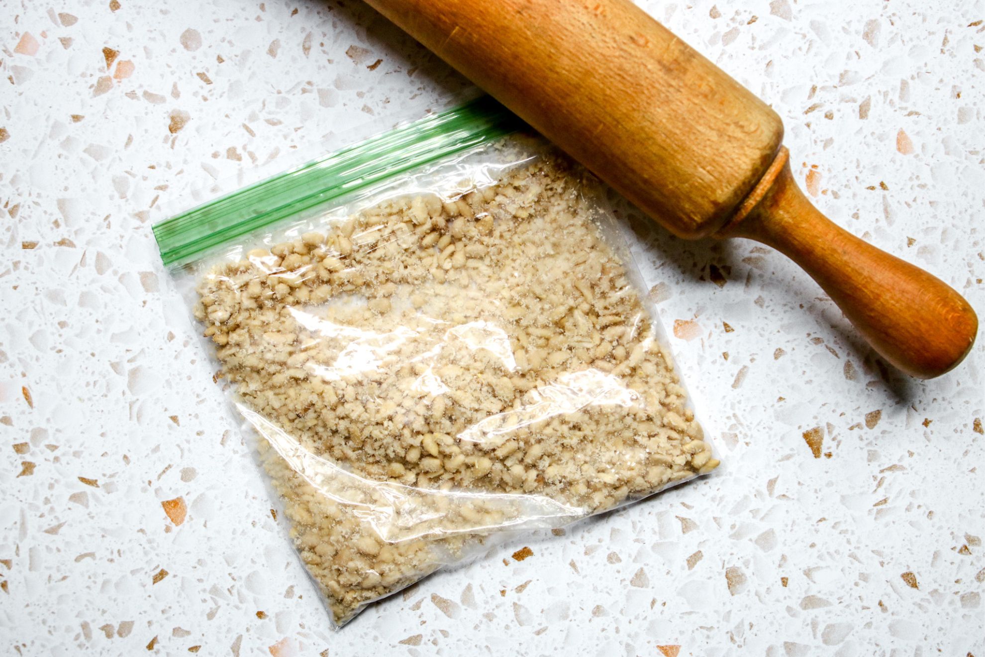 This is an overhead horizontal image of a small plastic baggie with crushed pine nuts in it. The baggie sits on a white terrazzo style counter. A rolling pin is on the counter above the baggie.