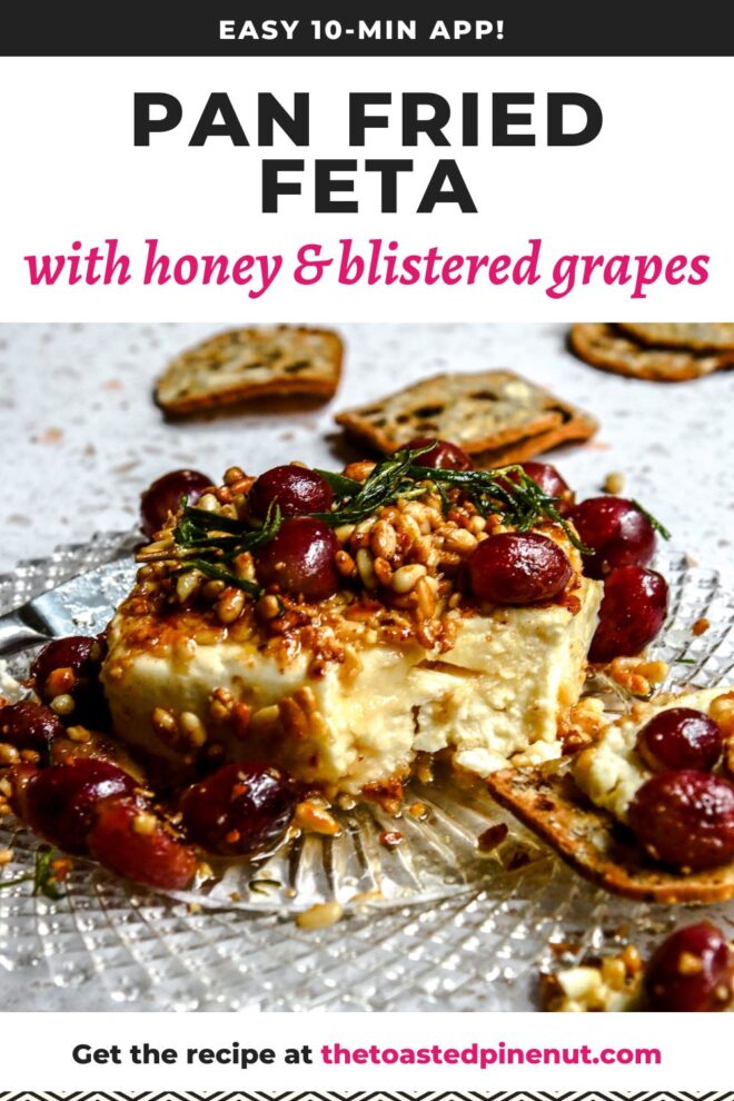 This is a vertical image looking at a block of feta from the side. The feta block is coated with toasted pine nuts. Blistered red grapes are on top of the feta and around it on the beveled glass plate. A sprig of rosemary is on top of the feta. Nutty crackers with dried fruit in them lay on the white terrazzo surface in front of and behind the plate. Text overlay reads "easy 10-min app! pan fried feta with honey & blistered grapes."