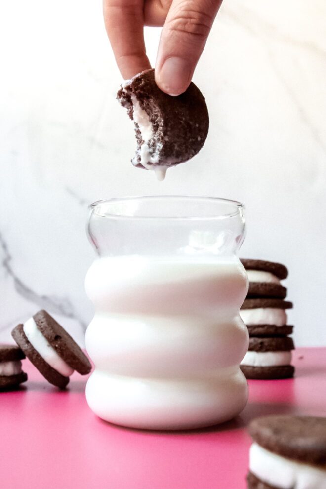This is a vertical image looking at a glass of milk from the side. The cup is filled 3/4 the way with milk and is sitting on a. pink surface with homemade oreos surrounding it. Coming from the top center of the image is a thumb and forefinger holding an oreo with a bite taken out of it. Milk is soaking the cookie sandwich and a drop of milk is hanging from the bottom of the cookie.