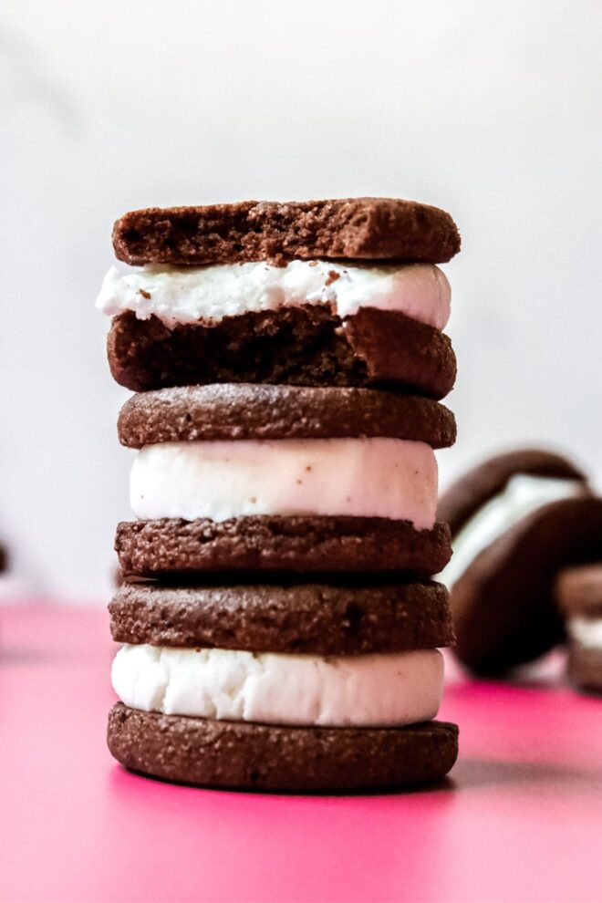 This is a closeup stack of three homemade oreos on a dark pink surface and a white background. More oreos are leaning on their side and blurred behind the stack. The top oreo has a bite taken out of it. T