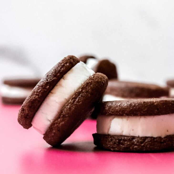 This is a vertical image of two oreos from a side view to reveal their layers. One oreo is leaning against the other oreo. Both oreos sit on a dark pink surface with more oreos blurred in the white marble background.