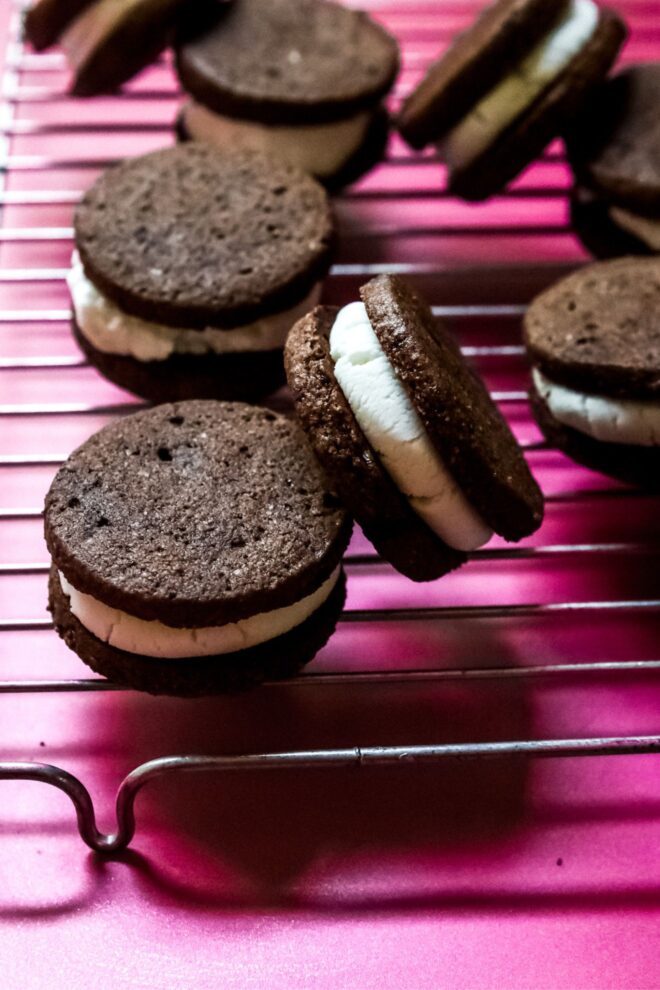 This is a vertical image looking at a a silver cooling rack from an angled view. The image looks onto chocolate sandwich cookies with a vanilla filling. Some cookies are flat on the cooling rack and some are leaning against the others.