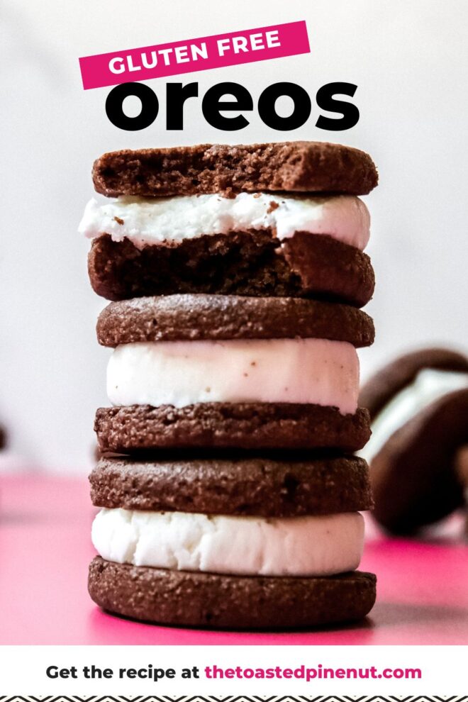 This is a closeup stack of three homemade oreos on a dark pink surface and a white background. More oreos are leaning on their side and blurred behind the stack. The top oreo has a bite taken out of it. Text overlay reads "gluten free oreos."