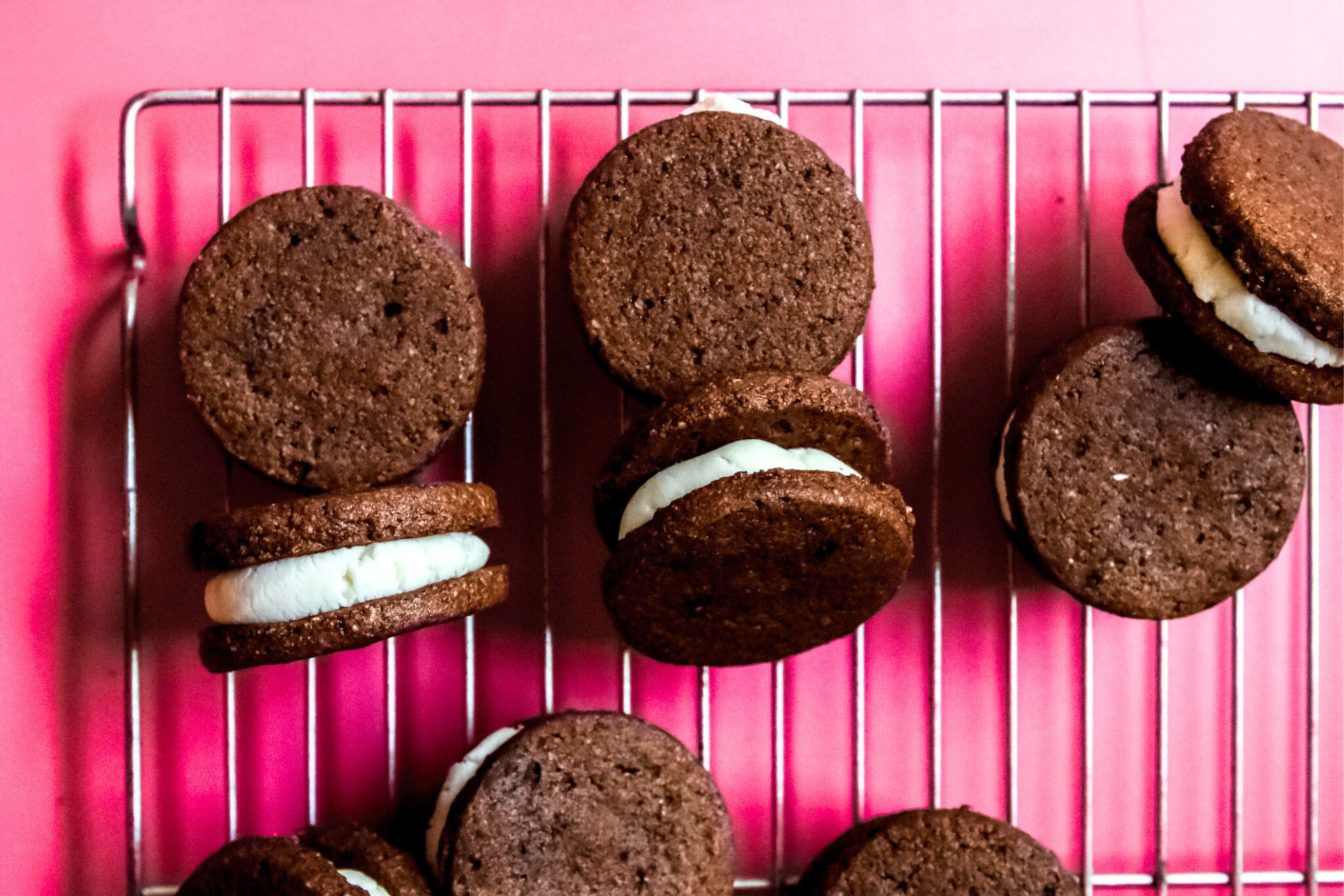 This is an overhead horizontal image of a silver cooling rack on a dark pink surface. On the cooling rack are chocolate sandwich cookies with vanilla icing in the middle. Some sandwich cookies are flat and some are leaning on their sides to reveal their layers.